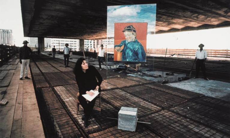 Lina Bo Bardi sul cantiere del costruendo MASP, da lei progettato tra il 1957 e il 1969, ritratta accanto ad uno dei cubi di cemento concepiti come sostegno dei pannelli di cristallo trasparente su cui appendere le opere d’arte esposte.